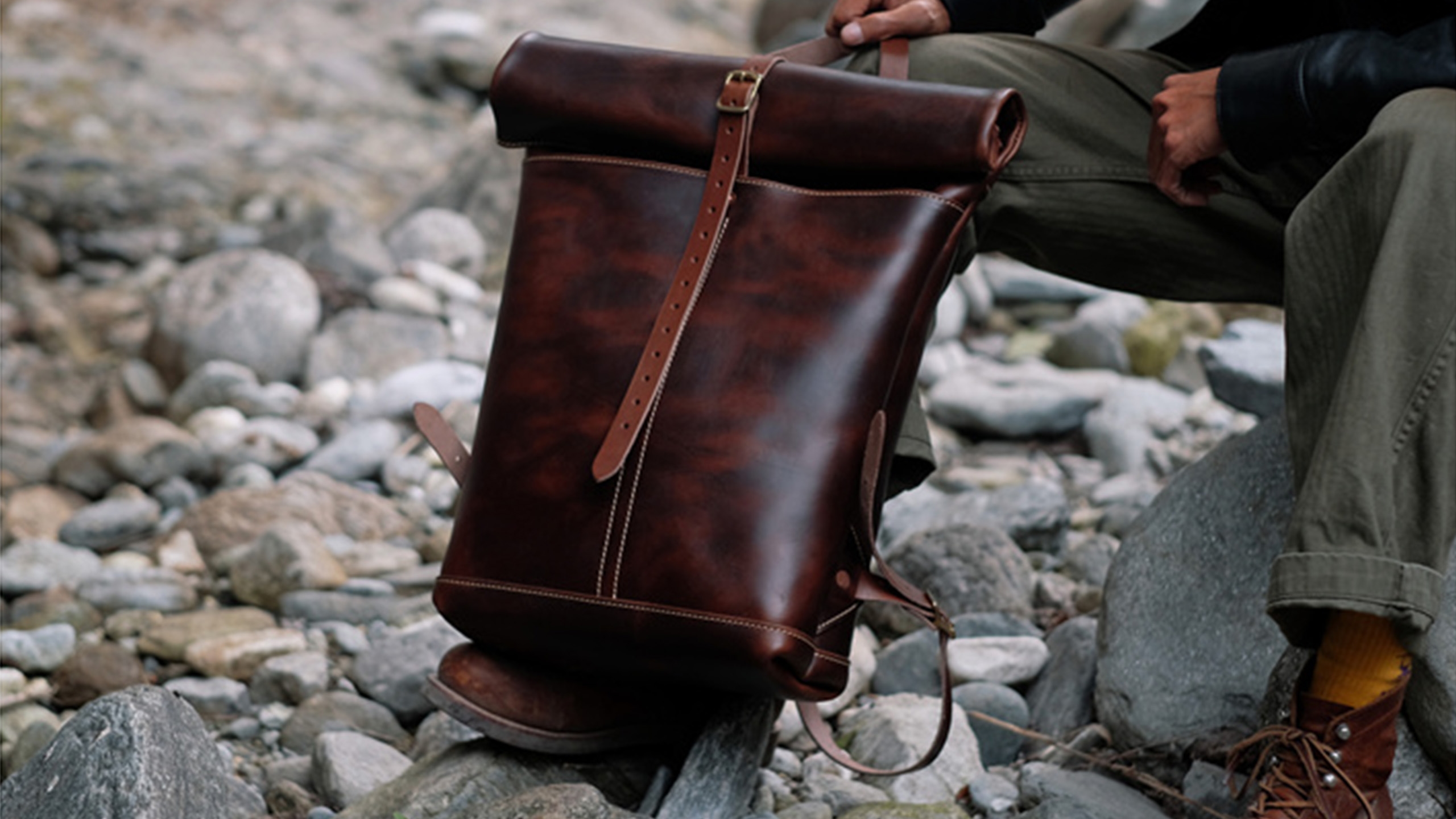 A dark leather rolltop backpack beside a rocky trail